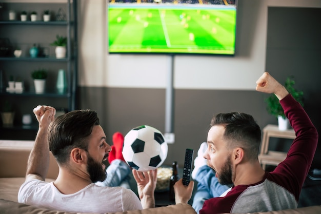 Vista traseira de dois melhores amigos e fãs de futebol assistindo a algum jogo esportivo na TV e bebendo cerveja e comendo lanches enquanto torcem pelo time no sofá