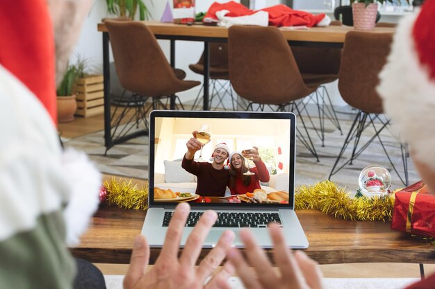 Foto vista traseira de homem e filho com chapéu de papai noel acenando enquanto faz uma videochamada com casal com chapéu de papai noel toa