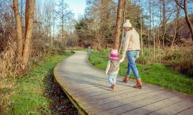 Vista traseira de mãe e filha caminhando juntas de mãos dadas sobre um caminho de madeira na floresta