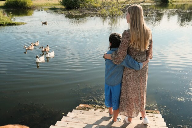 Vista traseira de mãe e filha se abraçando enquanto estão na ponte e observam os cisnes no rio ao ar livre