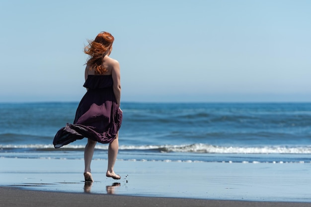 Vista traseira de mulher de vestido longo andando na praia de areia em direção à água no fundo do mar azul