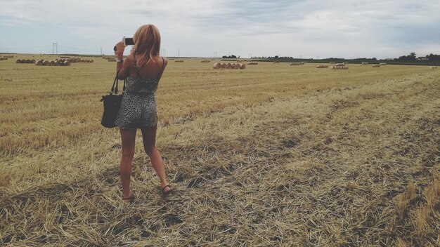 Foto vista traseira de mulher fotografando com telefone celular enquanto está de pé no campo contra o céu