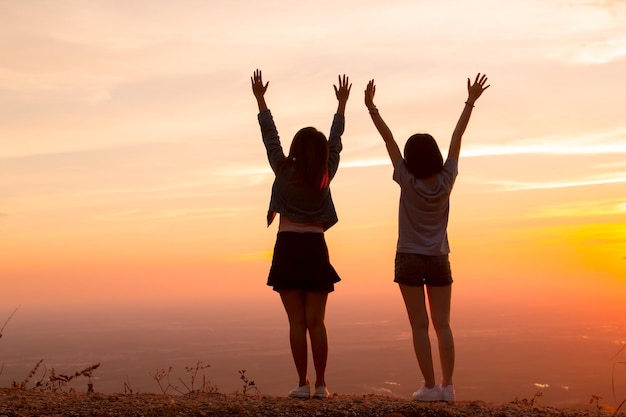 Vista traseira de mulheres com os braços levantados de pé contra o céu durante o pôr do sol
