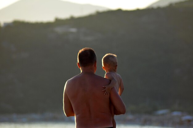 Foto vista traseira de pai sem camisa segurando filho enquanto está de pé ao ar livre