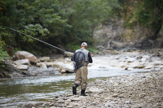 Vista traseira de pescador experiente vestido com roupas à prova d'água, caminhando ao longo do rio de montanha e pescando com vara. conceito de estilo de vida ativo.