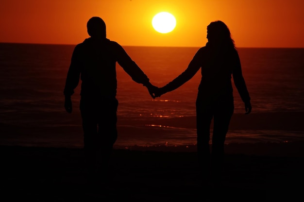 Foto vista traseira de um casal de silhuetas de mãos dadas na praia contra o céu durante o pôr do sol