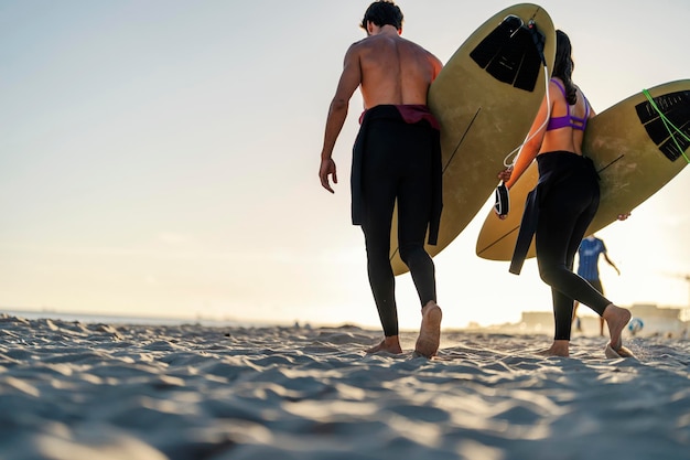 Vista traseira de um casal de surfistas carregando pranchas de surf na praia