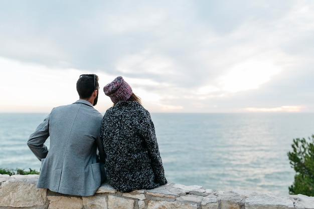 Vista traseira de um casal sentado em uma parede de contenção junto ao mar