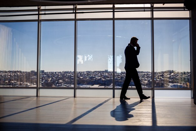 Vista traseira de um empresário, olhando por uma grande janela com vista para a cidade. Ele tem um telefone nas mãos. Visualização horizontal.