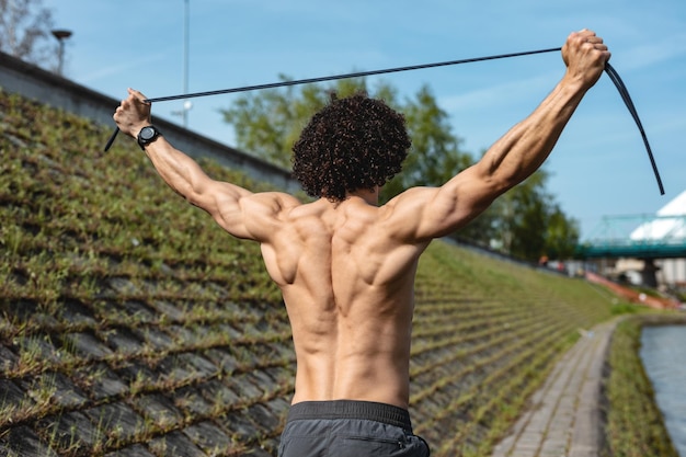 Vista traseira de um fisiculturista com torso nu, que está fazendo treinamento de corpo forte com elástico, perto do rio.