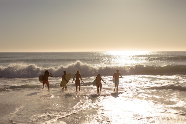 Vista traseira de um grupo multiétnico de amigos homens e mulheres de férias em uma praia segurando pranchas de surf, correndo para o mar em direção a uma onda enquanto o sol se põe