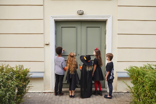 Vista traseira de um grupo multiétnico de crianças usando fantasias de halloween, tocando a campainha enquanto fazem doces ou travessuras juntos, copie o espaço