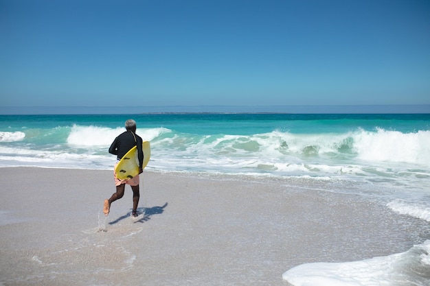 Vista traseira de um homem afro-americano sênior em uma praia ao sol, carregando uma prancha de surf sob o braço e correndo, com o céu azul e o mar ao fundo
