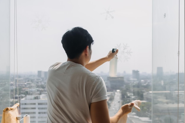 Vista traseira de um homem asiático tailandês limpando o vidro da janela em um quarto de apartamento com vista para a cidade. Mantenha o vidro.