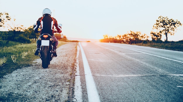 Foto vista traseira de um homem caucasiano bem equipado começando a andar de moto com um pôr do sol no fundo