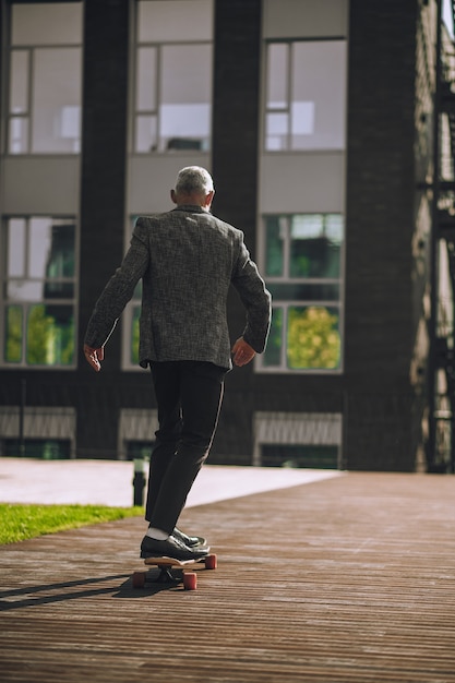 Vista traseira de um homem caucasiano de cabelos grisalhos andando de skate em frente ao prédio de escritórios