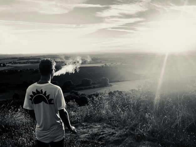 Foto vista traseira de um homem exhalando fumaça enquanto está de pé em uma colina olhando para a paisagem contra o céu durante o pôr do sol