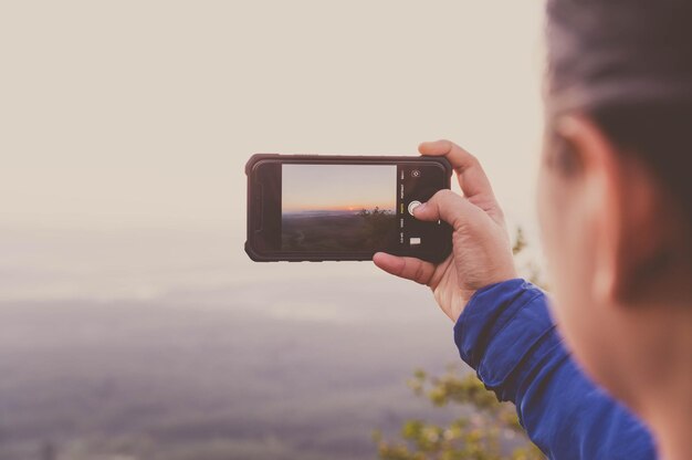 Vista traseira de um homem fotografando segurando um smartphone contra o céu