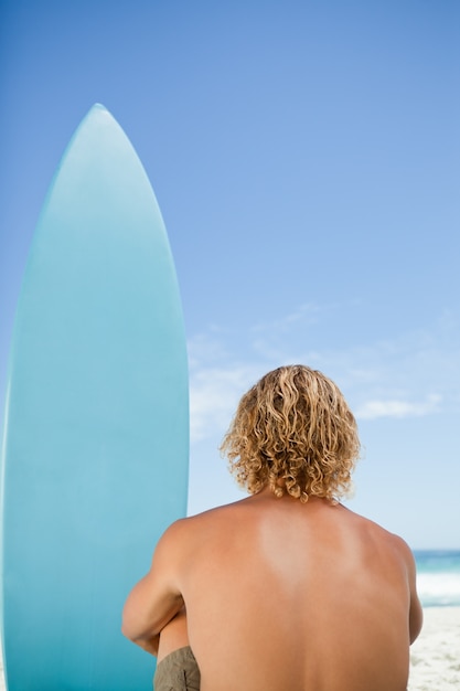 Vista traseira de um homem loiro olhando para o oceano sentado ao lado de sua prancha de surf