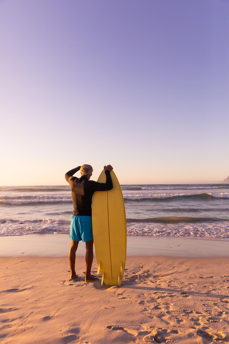 Foto vista traseira de um homem sênior afro-americano com uma prancha de surf olhando para a paisagem marinha contra um céu claro