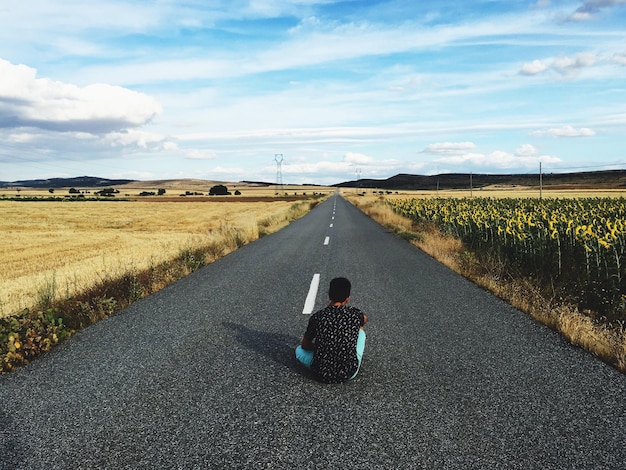 Vista traseira de um homem sentado na estrada no meio de um campo contra um céu nublado