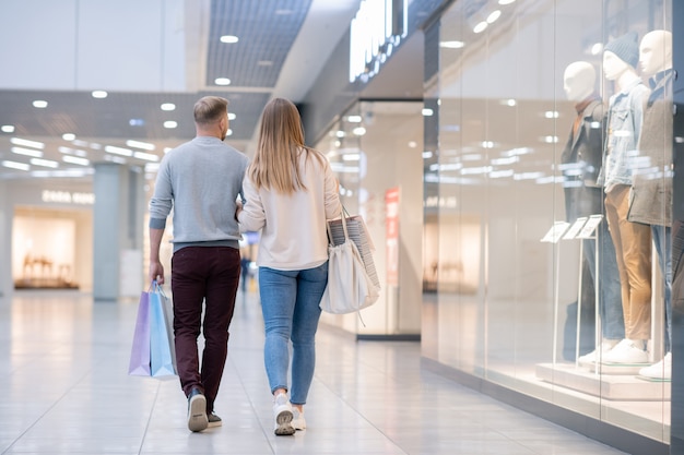 Vista traseira de um jovem casal casual se movendo ao longo da vitrine de uma loja no shopping enquanto a deixa após comprar o que quer