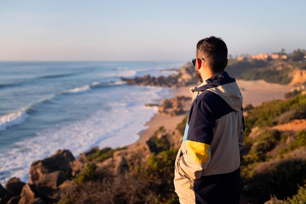 Vista traseira de um jovem com jaqueta e óculos de sol olhando para o mar de um penhasco ao lado da praia lazer e relaxar espaço de cópia de conceito para texto