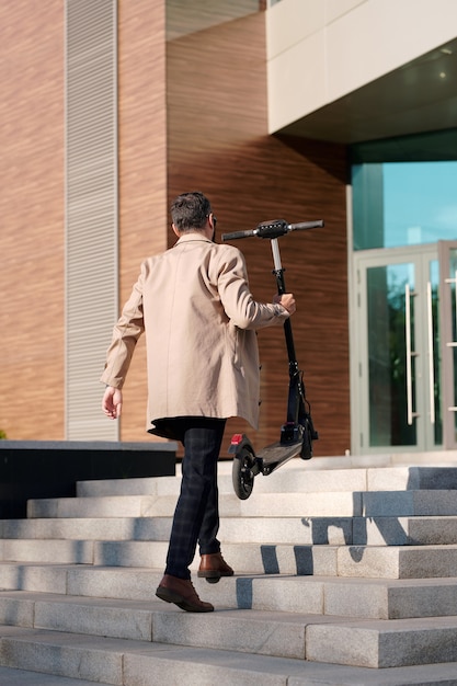 Vista traseira de um jovem empresário elegante em trajes casuais elegantes carregando uma scooter elétrica enquanto sobe as escadas para entrar no centro do escritório