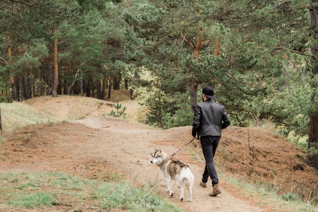 Foto vista traseira de um jovem segurando a coleira de um lindo cão husky de raça pura enquanto ambos relaxam em um ambiente rural