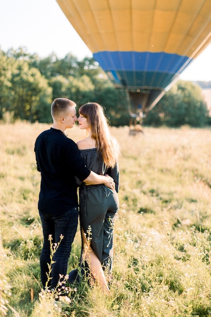 Vista traseira de um lindo casal romântico em roupas pretas elegantes, abraçando-se, em pé no campo ensolarado de verão com balão de ar quente