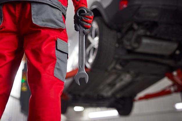 Vista traseira de um mecânico irreconhecível da colheita em uniforme e luvas em pé perto do carro com a chave inglesa na mão antes do processo de reparo na garagem