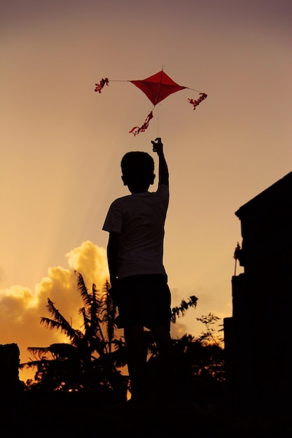 Vista traseira de um menino em silhueta voando com uma cometa contra o céu durante o pôr do sol