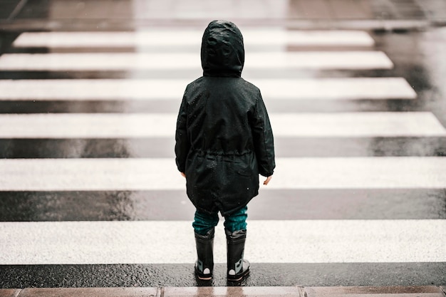 Vista traseira de um menino na capa de chuva e botas de pé na faixa de pedestres em dia chuvoso