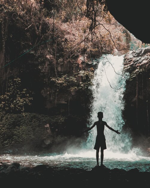 Foto vista traseira de um menino sem camisa de pé com os braços estendidos pela cachoeira