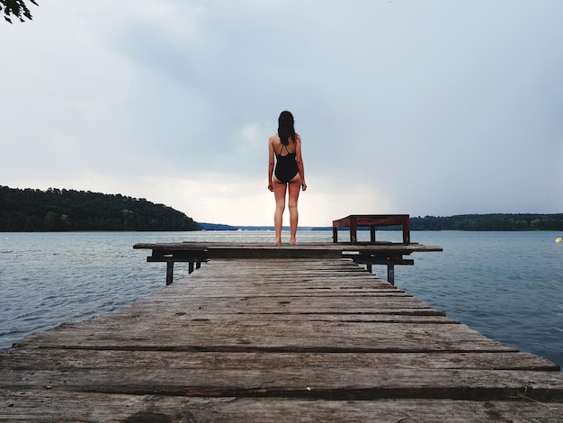 Foto vista traseira de uma adolescente de pé no cais sobre o lago contra o céu