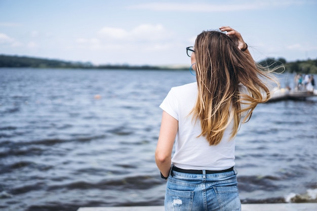 Vista traseira de uma bela jovem de óculos elegantes. Garota em camiseta branca posando no fundo da paisagem do lago