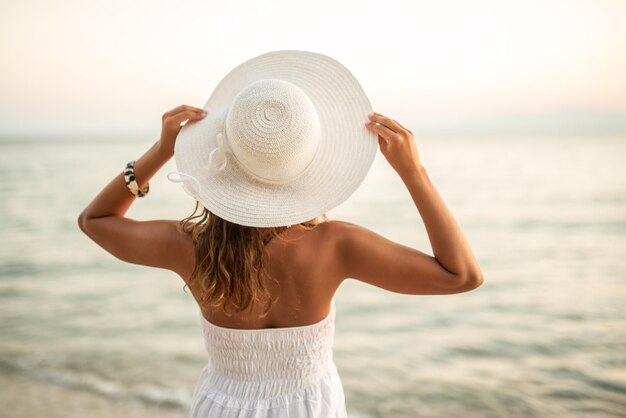 Vista traseira de uma bela jovem de vestido branco com chapéu na cabeça está relaxando e apreciando o pôr do sol na praia.