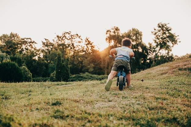 Vista traseira de uma criança em uma camiseta andando de bicicleta sozinha no campo de grama verde, à noite, pôr do sol
