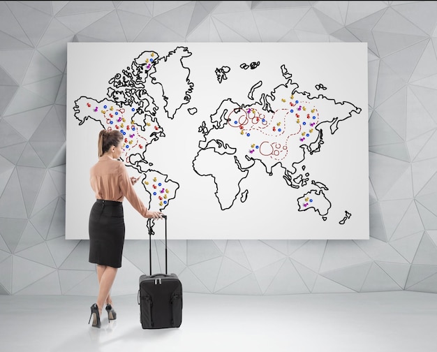 Vista traseira de uma empresária loira com uma mala preta em pé e olhando para um mapa-múndi com marcadores nele. Conceito de viagem. Elementos desta imagem fornecidos pela NASA