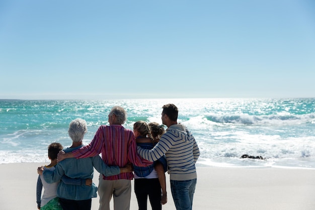 Vista traseira de uma família caucasiana de várias gerações de pé na praia com o céu azul e o mar ao fundo, abraçando e olhando para longe