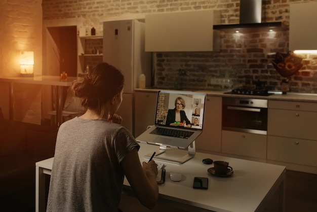 Vista traseira de uma funcionária que está trabalhando remotamente e anotando o discurso de um chefe durante uma videoconferência em um laptop de casa.