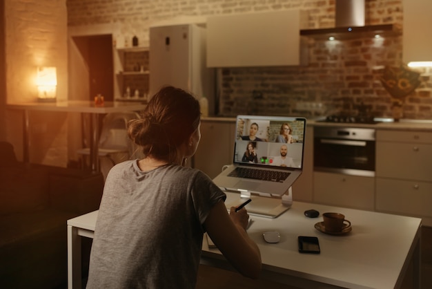 Vista traseira de uma funcionária que está trabalhando remotamente e conversando com seus colegas em uma videoconferência em um laptop de casa.