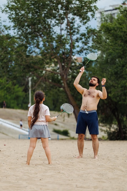 Vista traseira de uma garotinha bonita com longos cabelos escuros vestindo camiseta branca saia cinza jogando badminton com homem barbudo