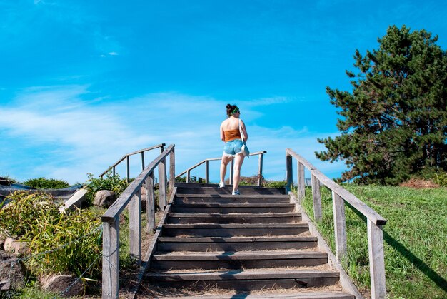 Vista traseira de uma jovem em shorts curtos subindo escadas em um parque de verão em um dia ensolarado.