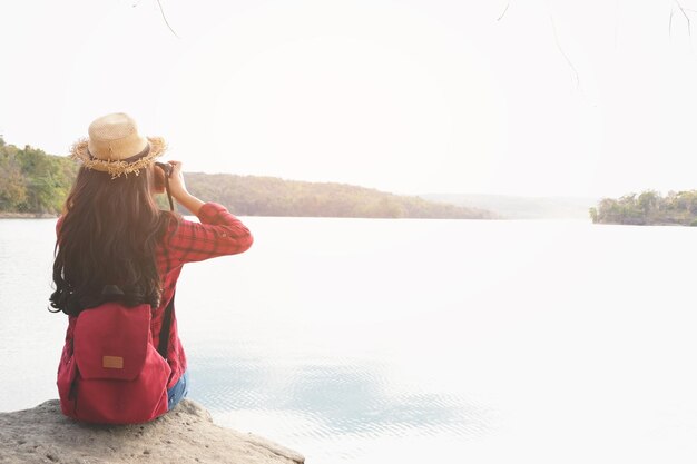 Foto vista traseira de uma jovem mulher com mochila sentada em uma rocha ao lado do lago contra o céu