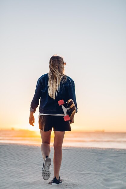 Vista traseira de uma jovem mulher com um skate caminhando na praia contra um céu claro durante o pôr do sol