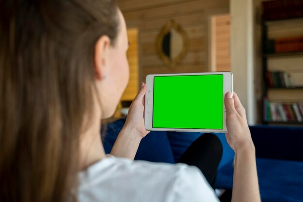 Vista traseira de uma jovem mulher segurando um tablet horizontal com tela verde sentada na sala