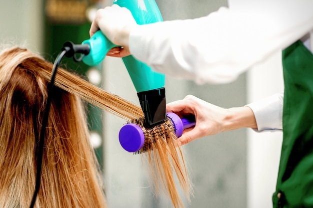 Vista traseira de uma jovem recebendo secando o cabelo com um secador de cabelo e uma escova de cabelo em um salão de cabeleireiro