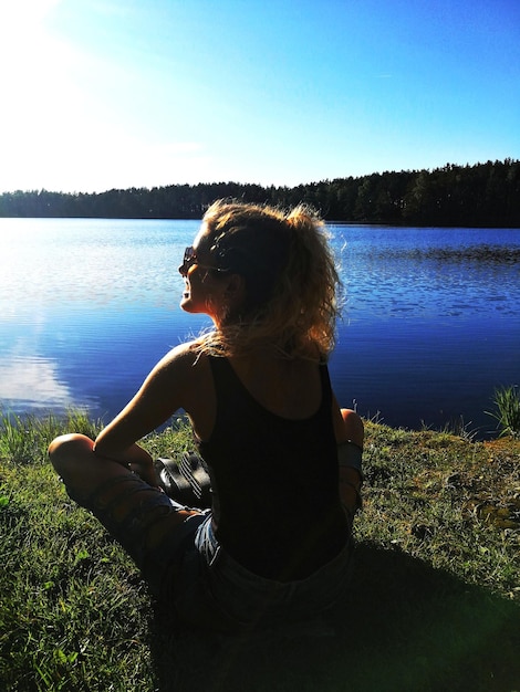 Foto vista traseira de uma jovem sentada ao lado do lago contra o céu azul durante um dia ensolarado