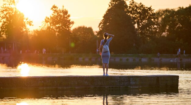 Vista traseira de uma jovem solitária em pé sozinho na margem do lago, desfrutando de uma noite quente. Bem-estar e relaxamento no conceito de natureza.